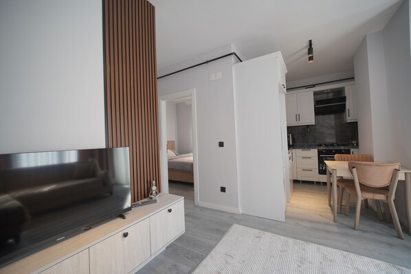A modern kitchen with a dining area, a TV on a wooden stand, and a visible bed in the adjacent room.
