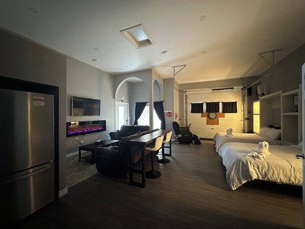 A modern hotel room with a flat-screen TV, a fireplace, a dining area with a table and chairs, and two beds.