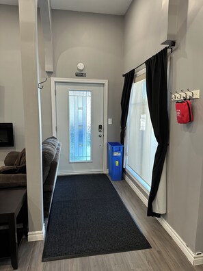 A hallway with a door leading to a balcony, a blue trash bin, a coat rack with a red bag, and a black doormat. - Modern Coach House (Cochrane)