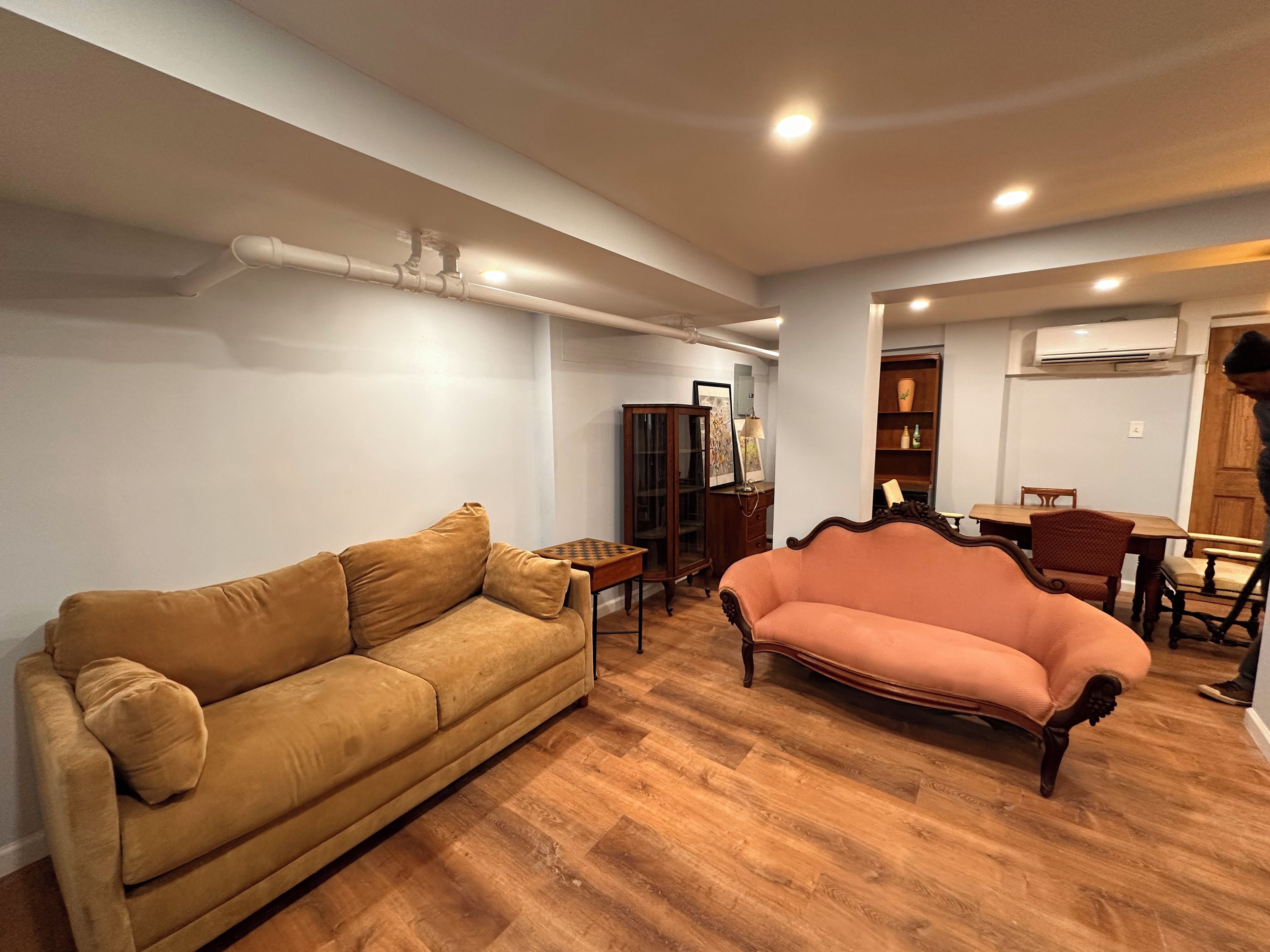 A living room with two sofas, a wooden herringbone floor, a dining area with a table and chairs, and an air conditioning unit on the wall.