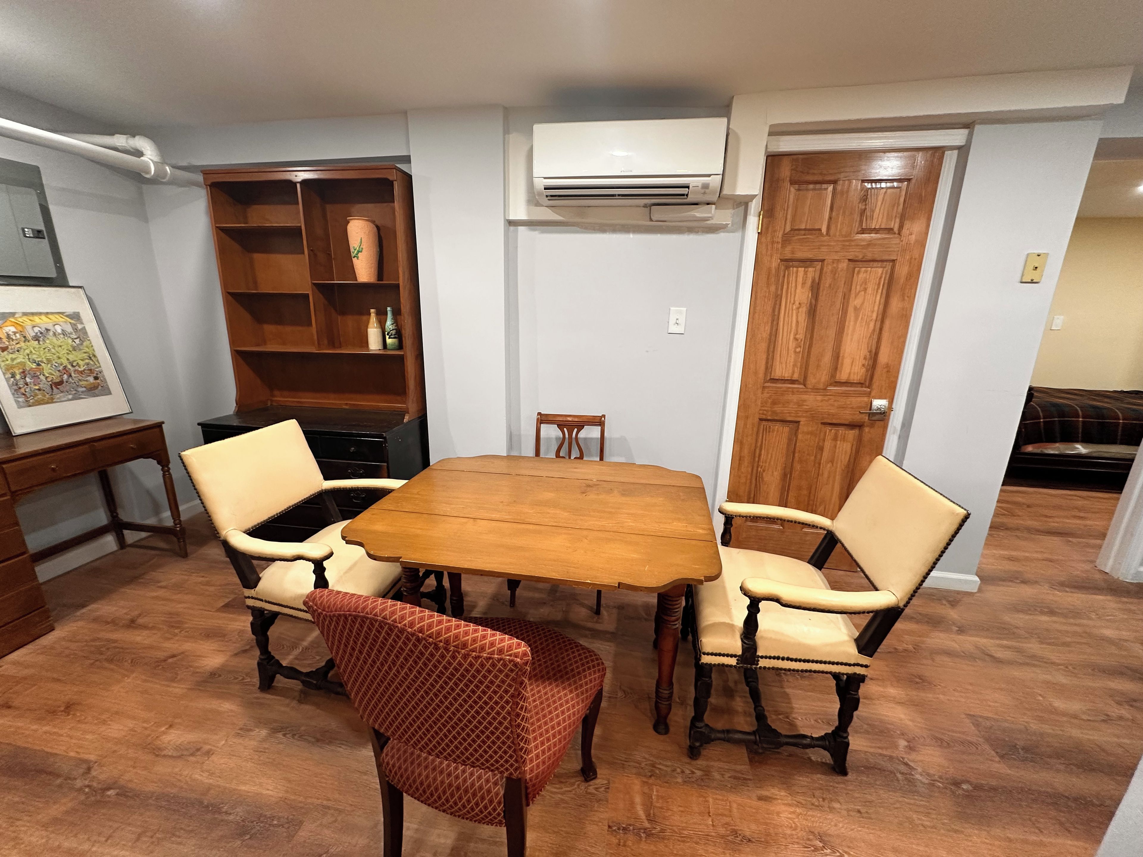 A dining room with a wooden table and four chairs, a bookshelf, and an air conditioning unit.
