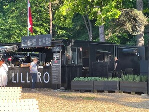 Un petit kiosque de vente de nourriture en plein air, avec un panneau affichant le menu, une pancarte indiquant « RETIREZ VOS COMMANDES ICI » et une autre pancarte indiquant « SERVICE BRUG ».