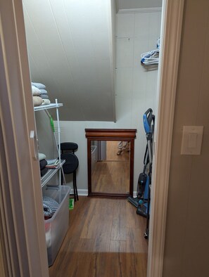 A narrow hallway with a wooden floor, a mirror, a vacuum cleaner, and a shelf with folded towels. - Cozy Flat, Super Central Location (Pensacola)