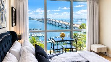 A bedroom with a large window overlooking a marina and a bridge, a bed with white and black bedding, a small table with a globe, and a chair.