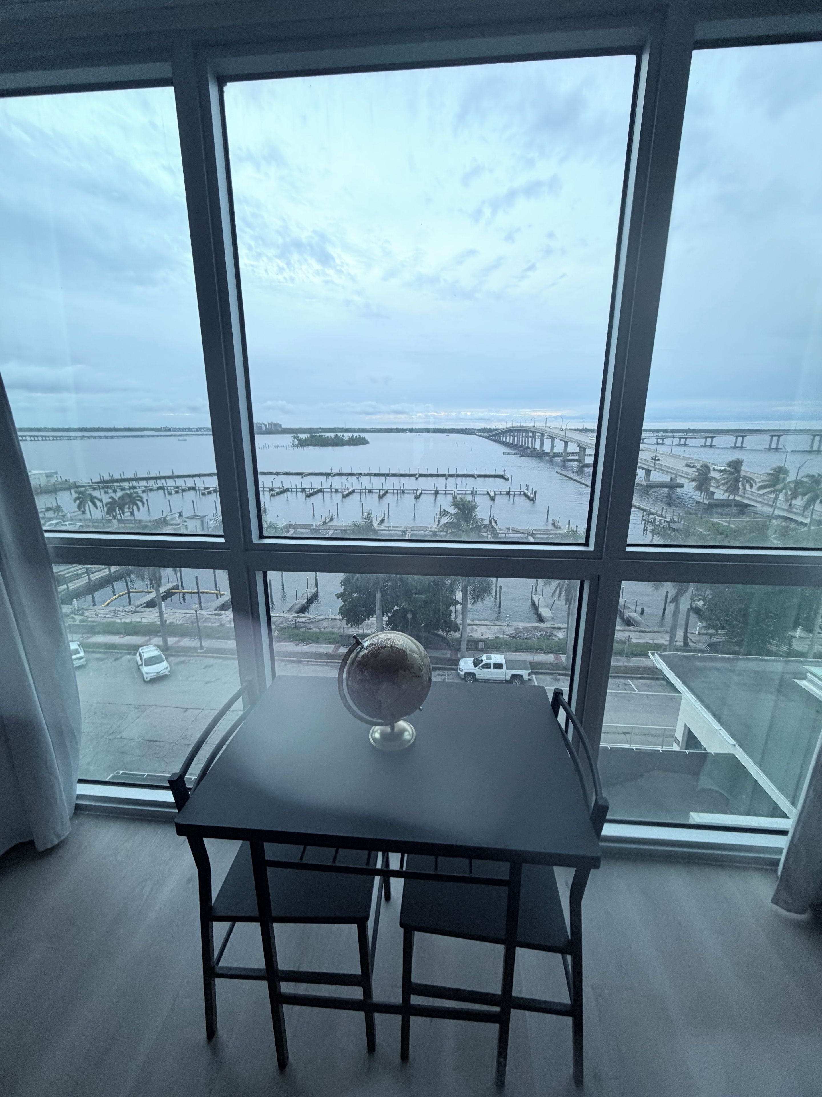 A room with a large window overlooking a marina and a bridge. There is a table with two chairs and a globe on it.