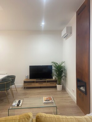 Ein Wohnzimmer mit Flachbildfernseher auf Holzfuß, Glastisch, gelbes Sofa, Topfpflanze und Holzschrank mit TV-Regal.