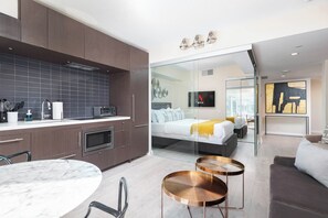 A modern hotel room with a kitchen area, a bed with a glass partition, and a dining area with a round table. - Luxury Yorkville 2BR, Panoramic Skyline Views (Toronto)