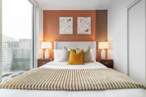A hotel room with a large bed, two bedside lamps, a view of the city, and two abstract paintings on the wall. - Downtown 3BR, Balcony & City Views (Toronto)