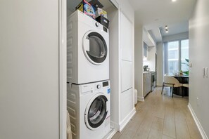 A laundry room with stacked washing machines and dryers, a kitchen area with a dining table and chairs, and a view through a window. - Downtown 3BR, Balcony & City Views (Toronto)