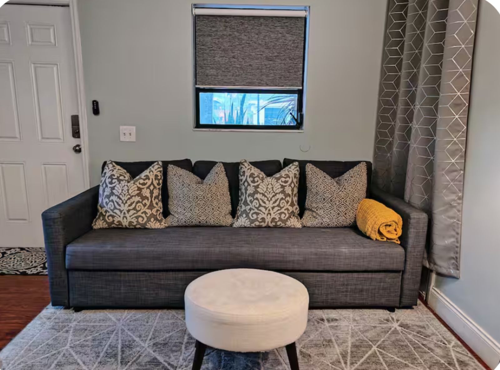 A grey sofa with patterned cushions, a small round ottoman, and a window with a view of trees.