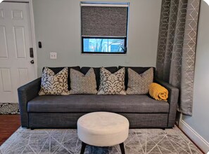 A grey sofa with patterned cushions, a small round ottoman, and a window with a view of trees. - Private King Suite 4-Min Walk to Wilton Drive (Wilton Manors)