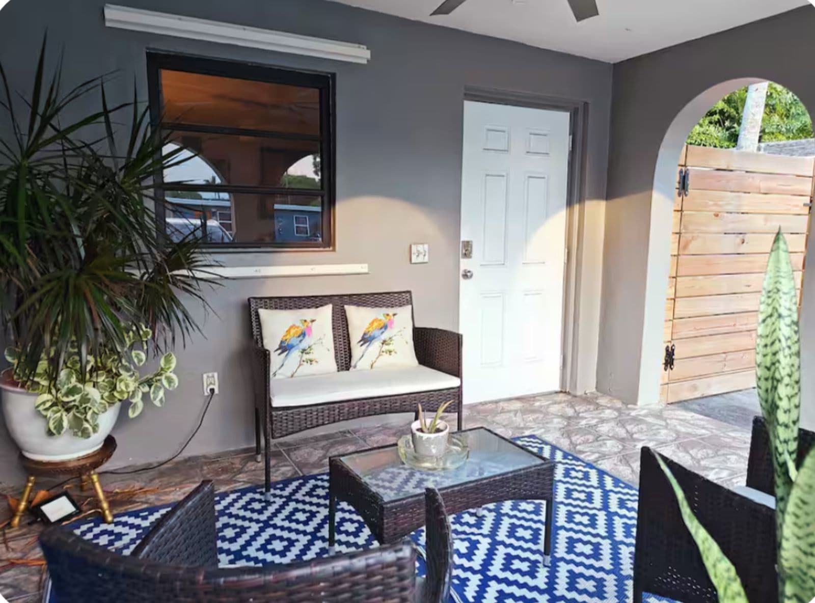 A room with a wicker sofa, a glass coffee table, a potted plant, and a view of a door and gate.