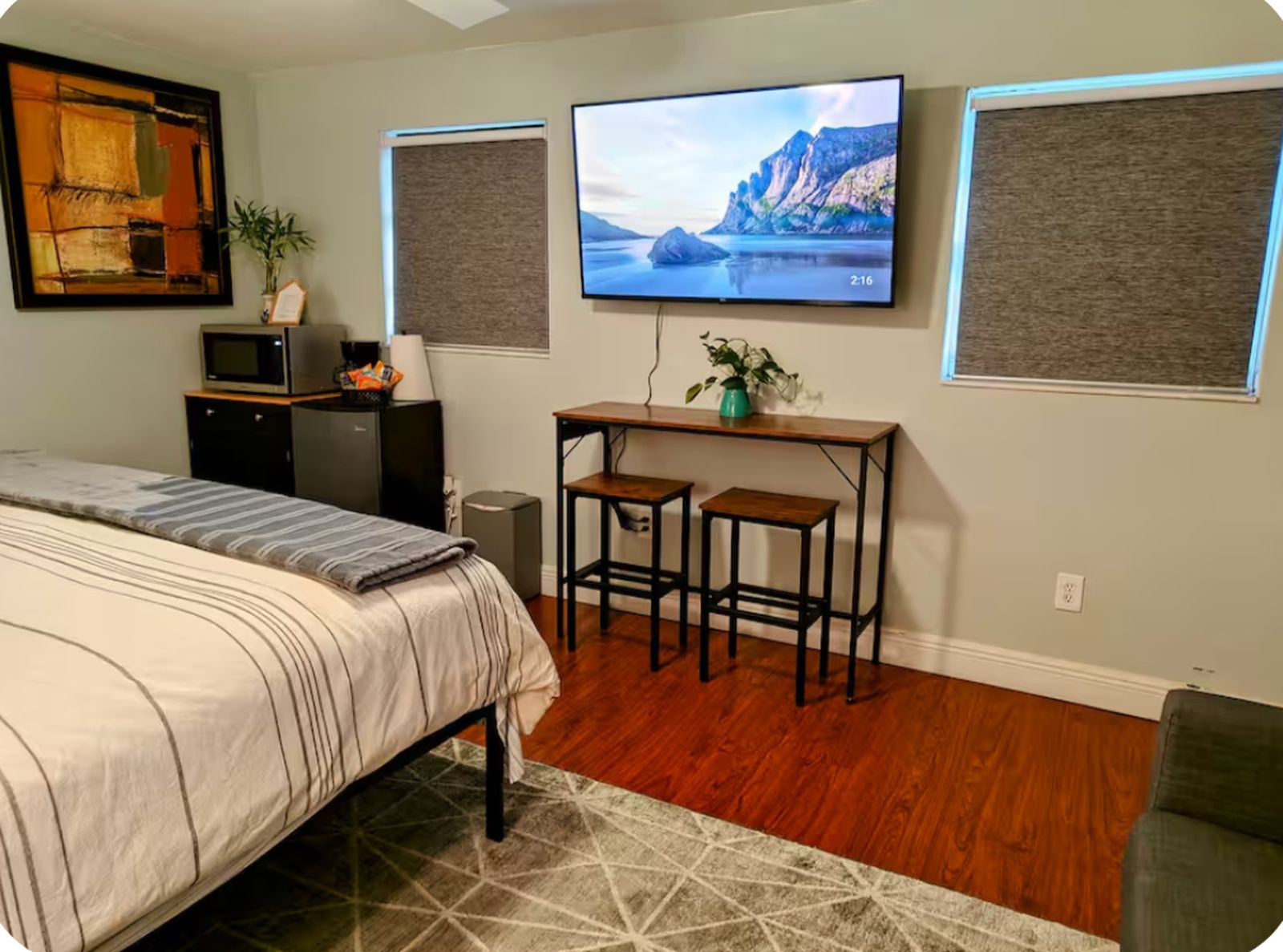 A bedroom with a bed, a desk with two stools, a television, and a wall-mounted speaker.