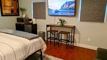 A bedroom with a bed, a desk with two stools, a television, and a wall-mounted speaker.