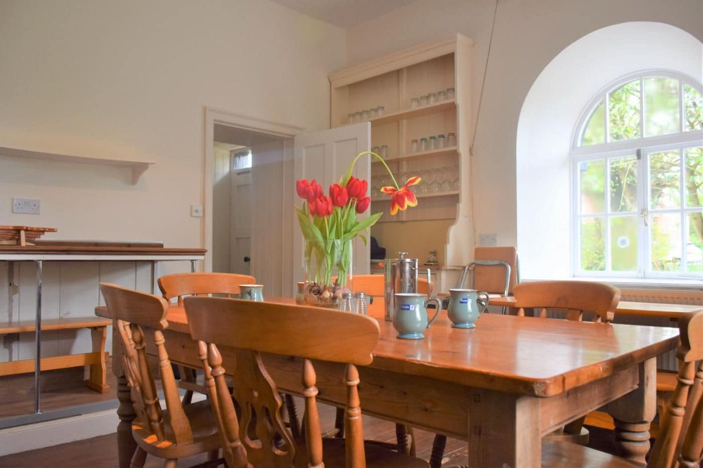 Un coin repas avec une table et des chaises en bois, un vase de tulipes rouges et une fenêtre donnant sur un paysage verdoyant.