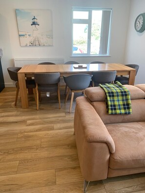 A dining area with a wooden table and chairs, a sofa with a plaid blanket, a painting of a lighthouse, and a clock on the wall. - Newly refurbished modern house with parking, ideally situated & sleeps up to 8! (Mevagissey)