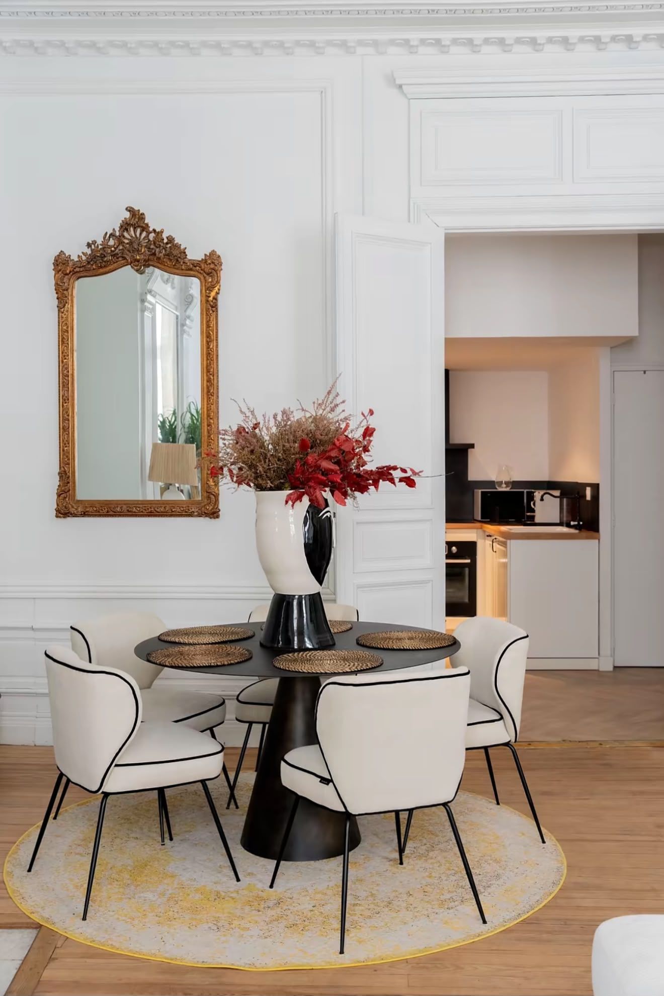 Een eetruimte met een ronde tafel, zes stoelen, een vaas met bloemen, een spiegel en een keuken op de achtergrond.