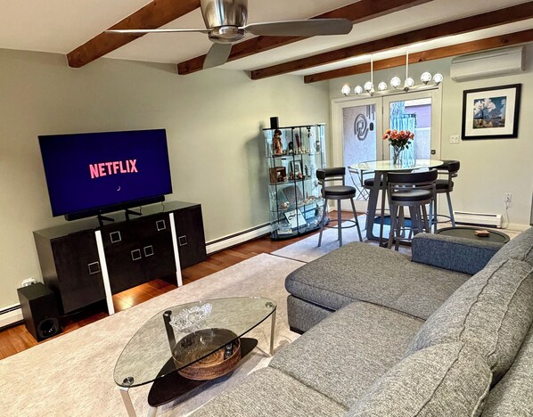 Ein Wohnzimmer mit einem Flachbildfernseher, der 'NETFLIX' anzeigt, einem Glastisch, einem grauen Sofa, einem Essbereich mit Stühlen und einer Glastauschauhütte.