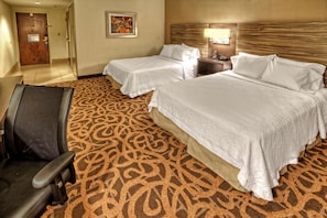 Quarto de hotel com duas camas, uma escrivaninha, uma cadeira e um tapete estampado.
