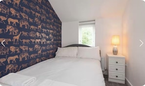 Ein Schlafzimmer mit einem Bett, weißer Bettwäsche, einem Nachttisch mit Lampe und einem Fenster mit Jalousien.