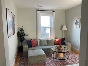 A living room with a sectional sofa, ottoman, glass coffee table, and a potted plant. - Boston Urban Nests U4: Long Term 1 Month + (Boston)
