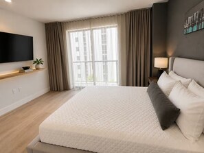 A modern hotel room with a large bed, a flat-screen TV, a bedside table with a lamp, and a view of buildings through the window. - Enjoyable 1-bedroom apartment in peaceful Miami neighborhood (Miami)