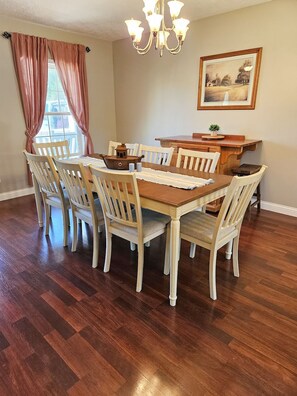 Uma sala de jantar com mesa e cadeiras de madeira, um lustre e um quadro na parede.