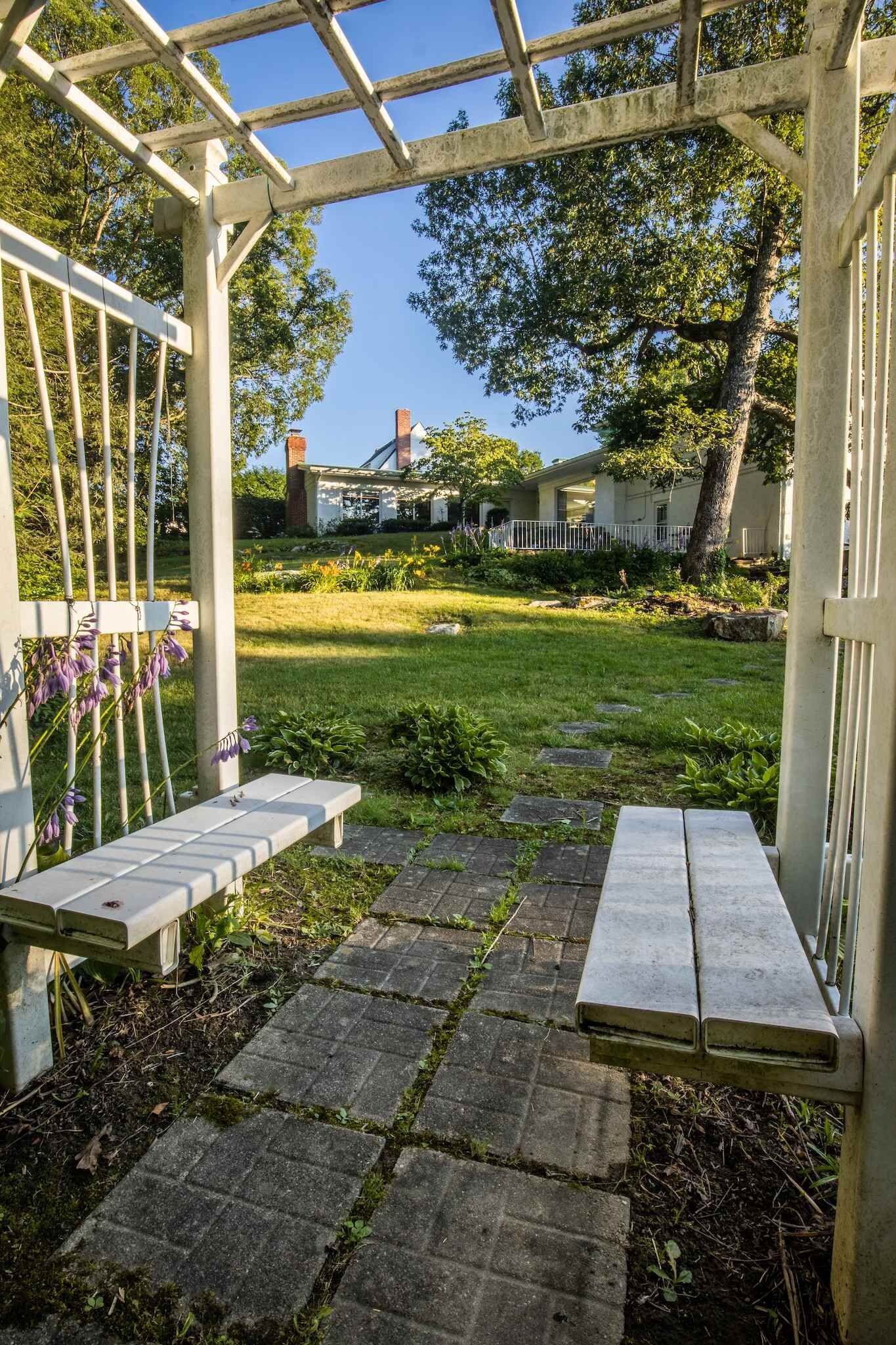 Een tuin met een pergola, twee bankjes en een geplaveid pad.