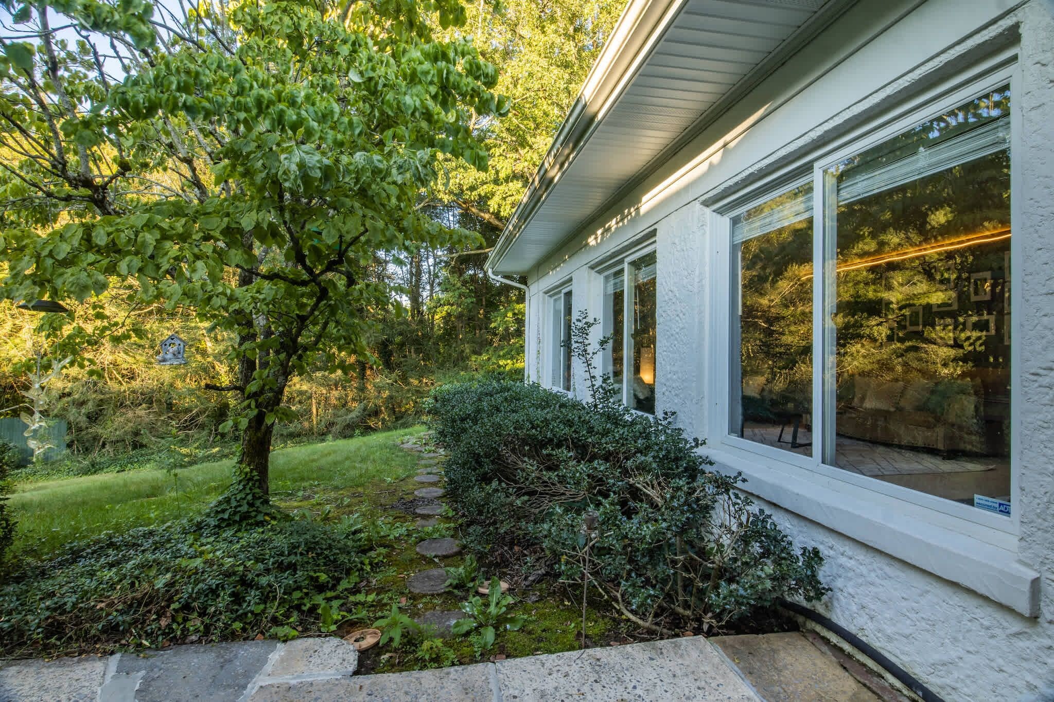A house with a white exterior, large windows, and a garden with a tree and shrubs.