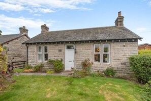 A stone cottage with a white door, two windows, and a small garden with flowers and shrubs. - 2 Bed in Winchburgh (oc-h44720) (Broxburn)