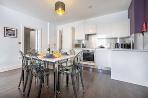 A modern kitchen with white cabinets, a stainless steel range hood, and a dining area with a glass table and chairs. - Stylish 2BR | Near City Centre | FREE Parking (Reading)