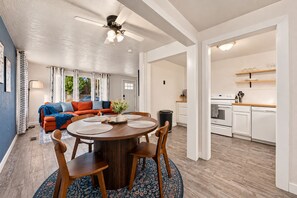 Een moderne keuken met witte apparaten, een eetgedeelte met een ronde tafel en stoelen, en een woonkamer met een bank en gordijnen.