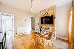 Ein modernes Wohnzimmer mit Essbereich, Holzböden, einem Glastisch und einem an der Wand befestigten Fernseher, der 'NETFLIX' zeigt.
