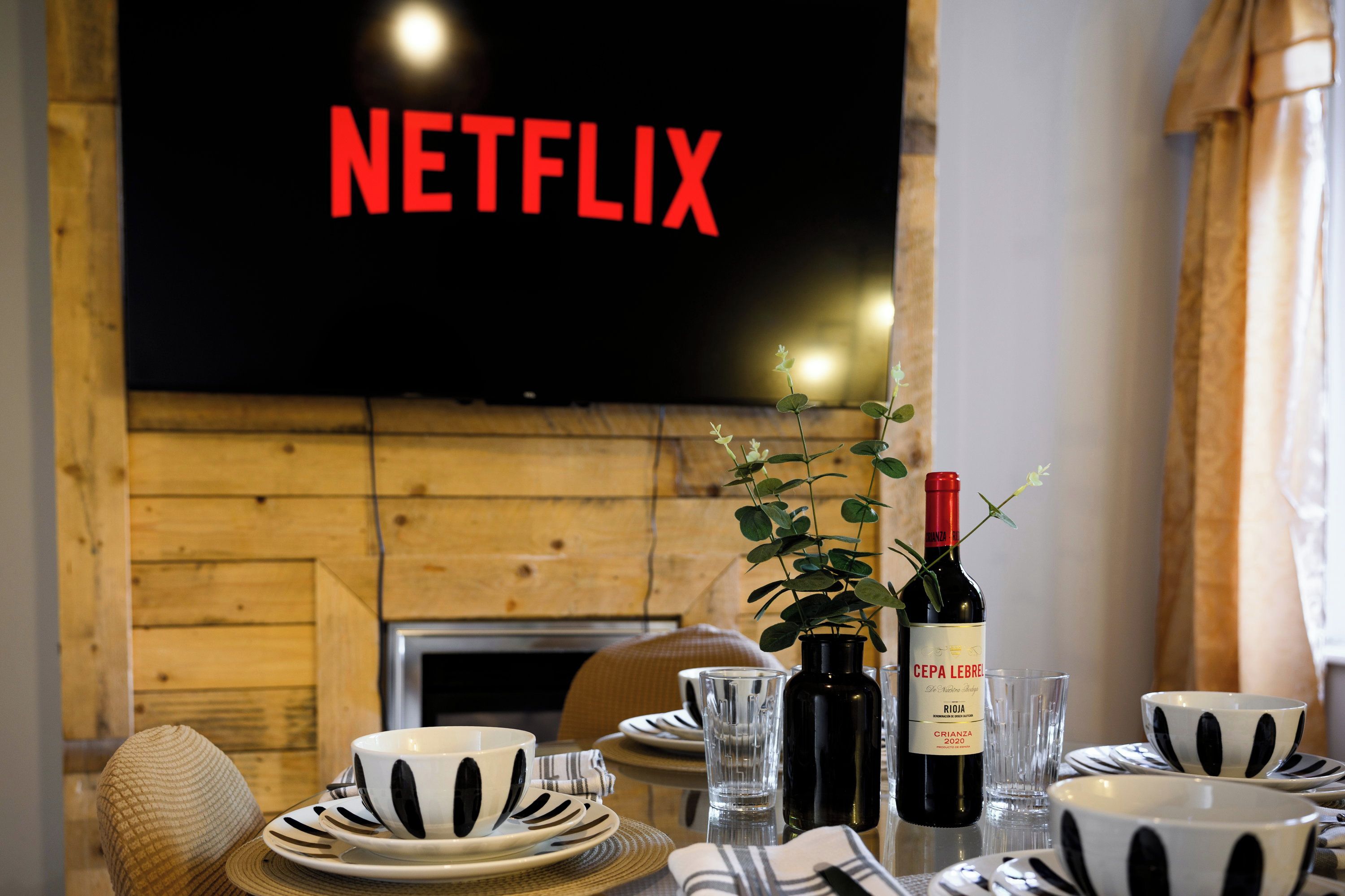 Ein Essbereich mit einem an der Wand montierten Netflix-Fernseher, einer Weinflasche und schwarz-weiß gemustertem Geschirr.