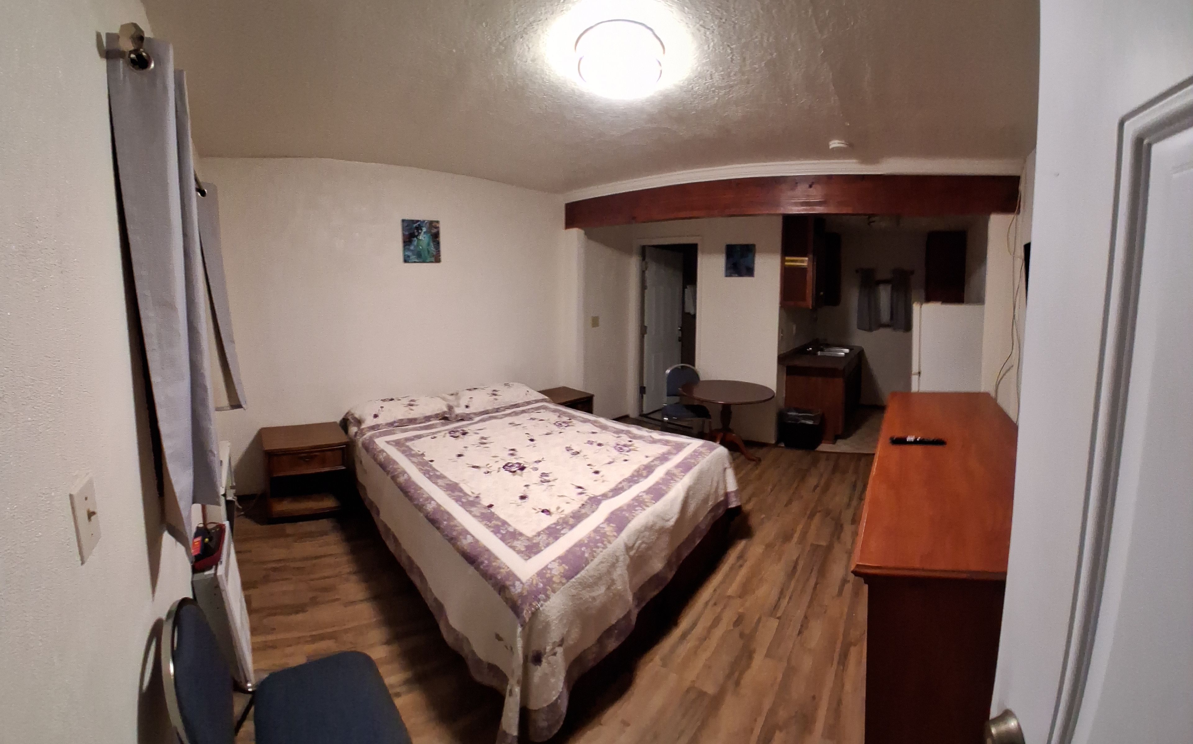 A compact hotel room with a bed, a small desk, a chair, and a kitchenette.