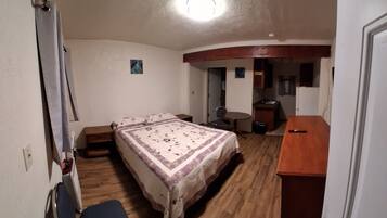 A compact hotel room with a bed, a small desk, a chair, and a kitchenette.