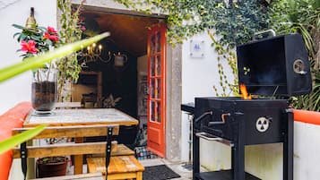 Une terrasse extérieure avec une table en bois, un banc, un barbecue et une porte donnant sur un espace cuisine.