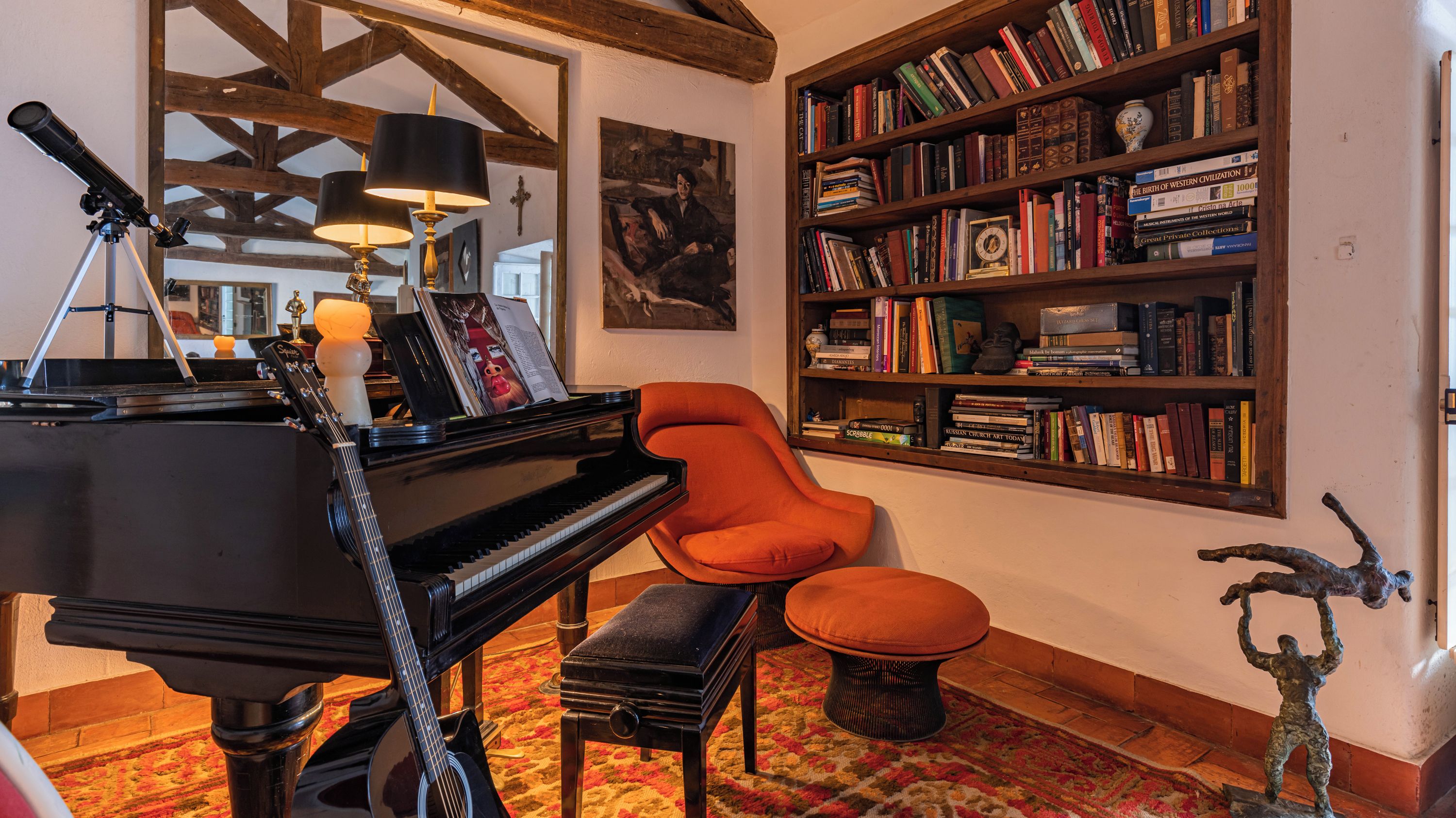 Ein Zimmer mit einem Flügel, einem Bücherregal voller Bücher, einem gemütlichen orangefarbenen Sessel und einer dekorativen Skulptur.
