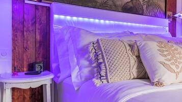 Una cama con sábanas blancas y almohadones decorativos, una mesita de noche con una lámpara, y una lámpara de pared.