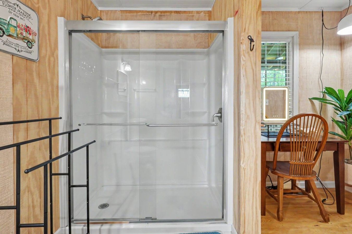A room with a glass shower enclosure, a wooden dining table with a chair, and a potted plant.