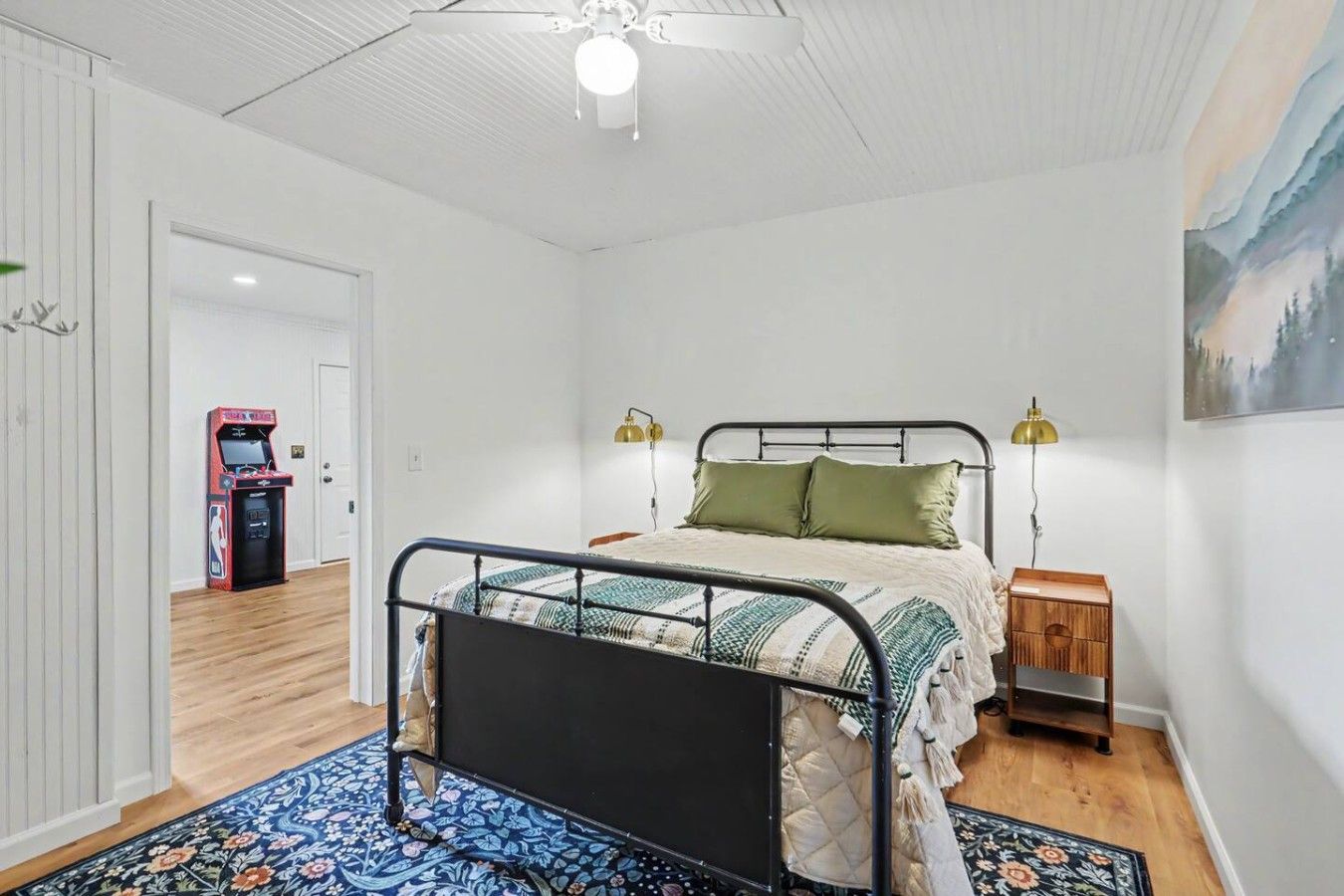 Una camera da letto con un letto, lampade sui comodini, un mobiletto, un armadio in legno, una ventola a soffitto e una porta che dà su un'altra stanza.