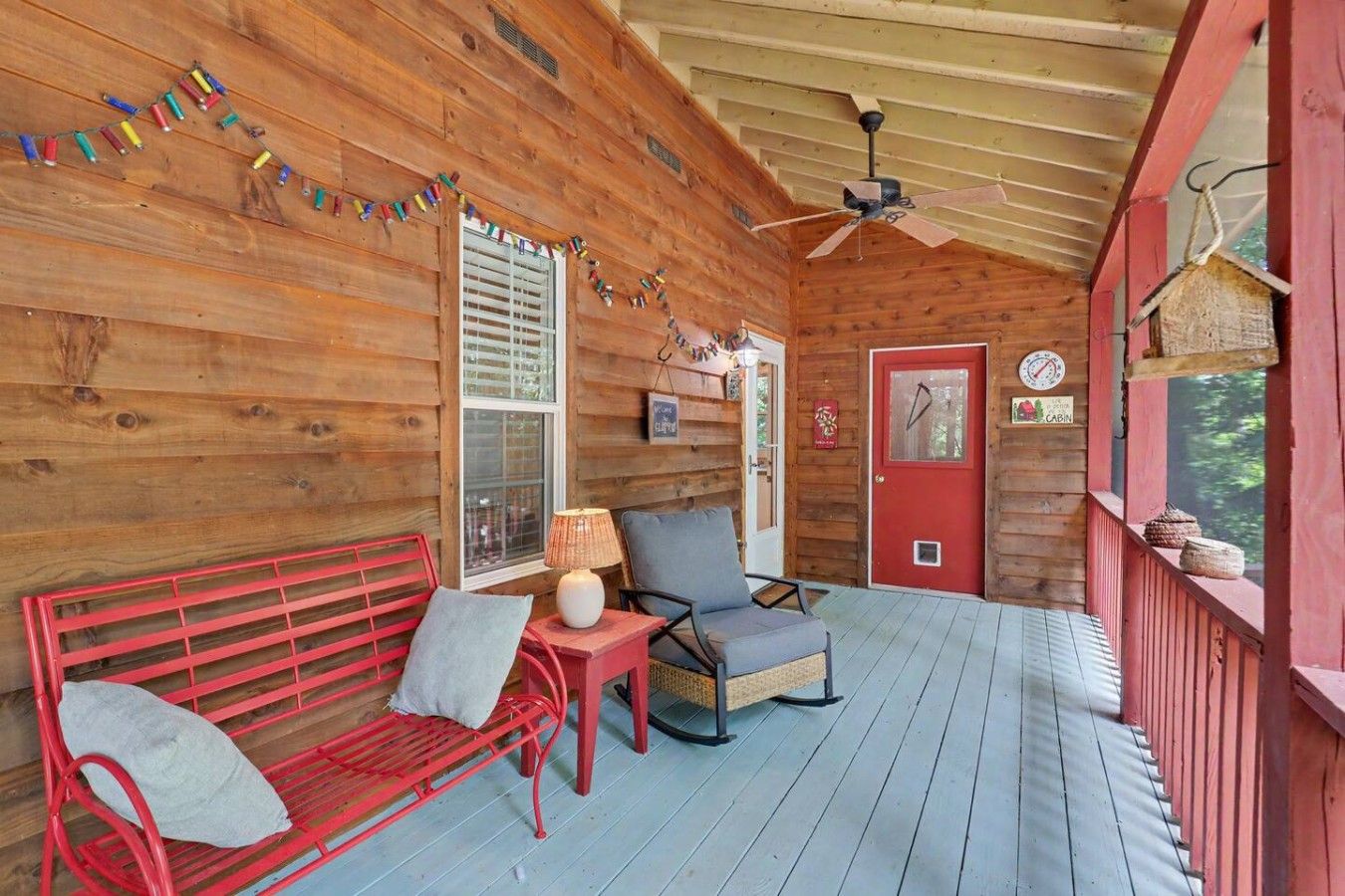 A wooden cabin with a porch, a red bench, a chair, a table with a lamp, and a red door.