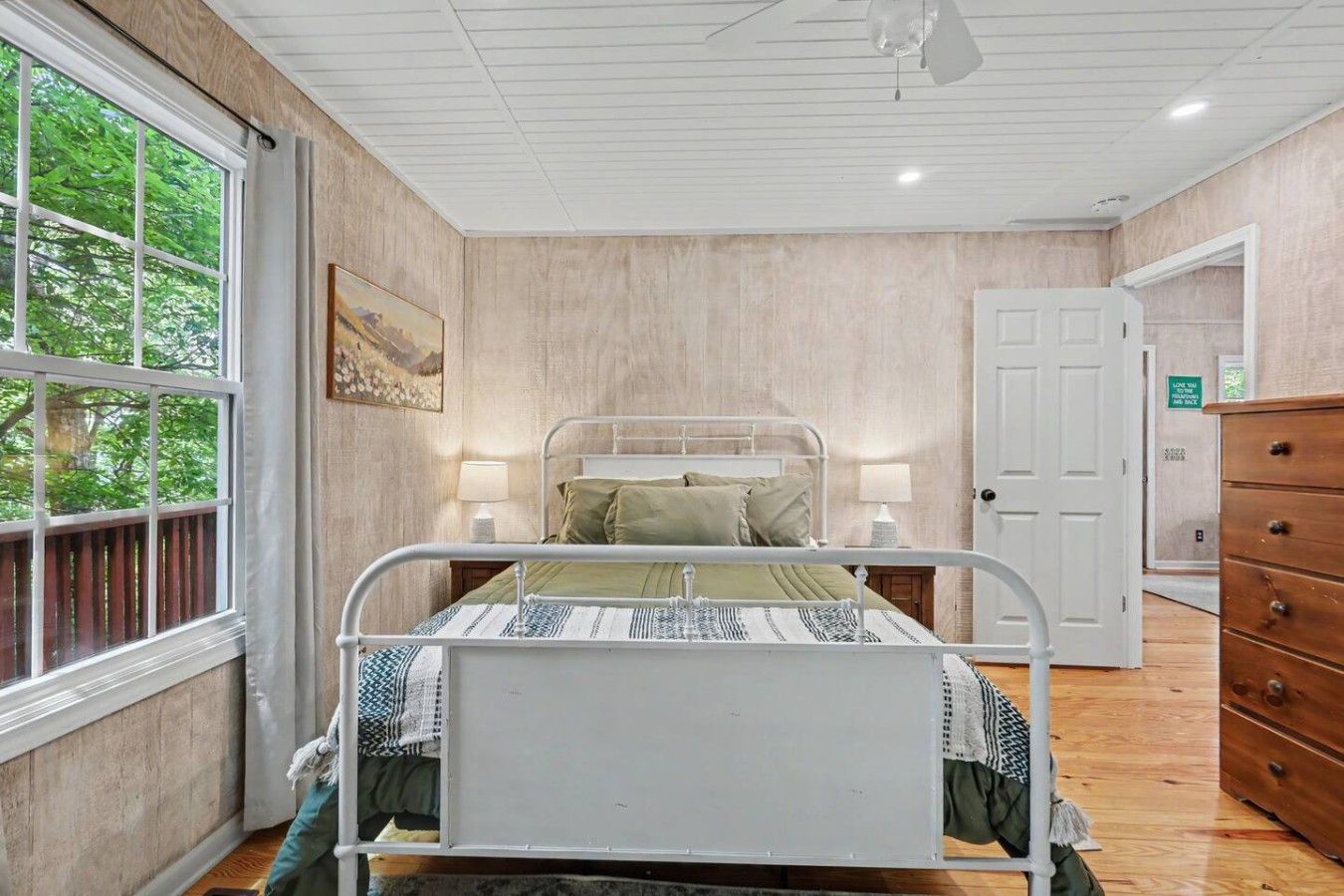 A bedroom with a metal bed frame, a wooden dresser, a door with a sign, and a window with a view of trees.