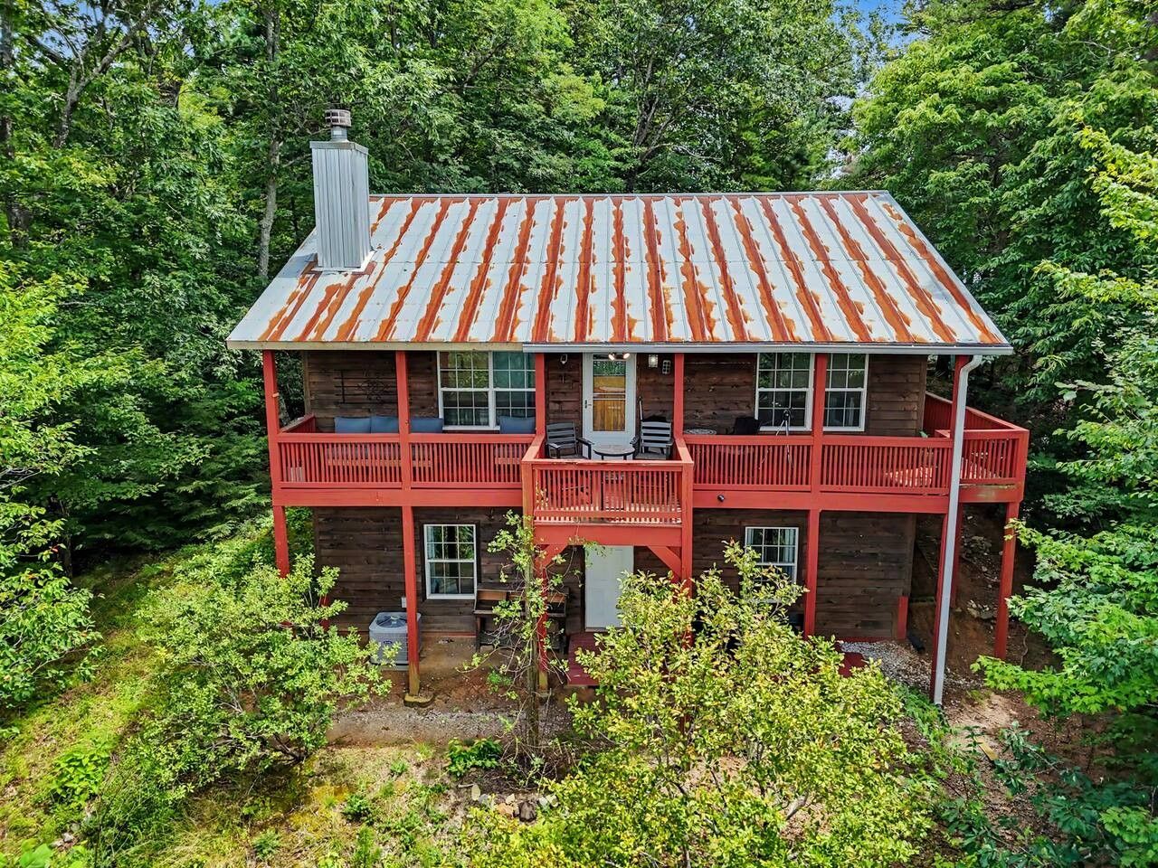 A two-story wooden cabin with a red balcony, surrounded by dense greenery.