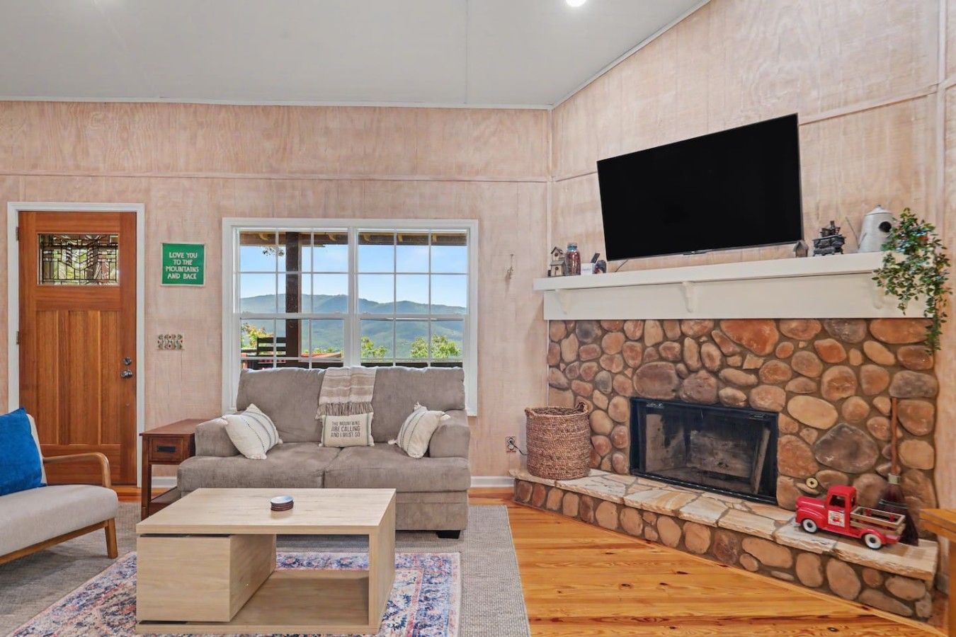 A living room with a stone fireplace, a flat-screen TV mounted above it, a sofa, a coffee table, and a door with a view of the mountains.