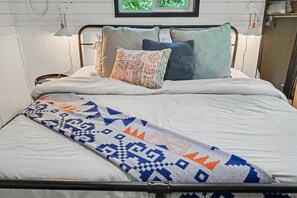 Cama com estrutura de metal, roupa de cama branca e manta decorativa estampada.