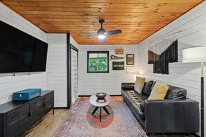 Ein Zimmer mit Holzbalkendecke, einem schwarzen Ledersofa, einem Fernseher, einem kleinen Tisch und einem gemusterten Teppich.