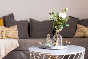 Una mesa redonda con un florero de vidrio que contiene flores blancas y hojas verdes, dos botellas de vidrio con tapones de corcho y una bandeja metálica con objetos decorativos.