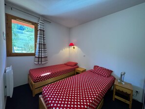 Ein kleines Zimmer mit zwei Einzelbetten, einem fensterbelaubten Fenster, einer Wandlampe und einer Heizung.