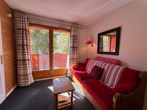 Een gezellige kamer met een rode bank, een klein houten tafeltje en een balkon met gordijnen.